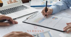 Overhead view of a financial meeting with two individuals analyzing charts and reports labeled 'Analysis Report' and 'Summary Sale,' alongside a laptop displaying graphs, representing cannabis business strategy planning by Verdant Strategies