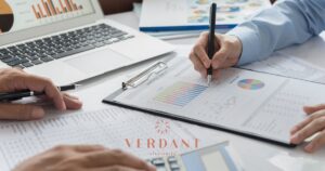 Overhead view of a financial meeting with two individuals analyzing charts and reports labeled 'Analysis Report' and 'Summary Sale,' alongside a laptop displaying graphs, representing cannabis business strategy planning by Verdant Strategies