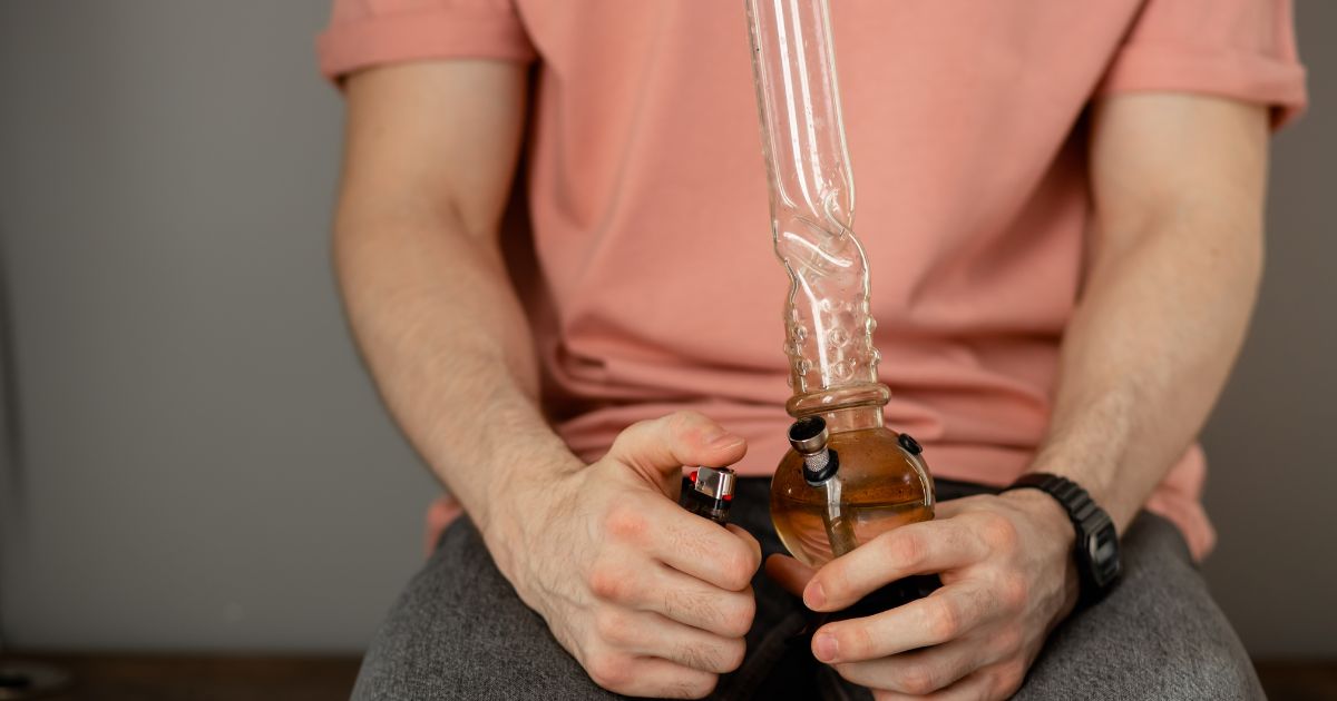 Close-up of a person holding a glass bong and lighter, wearing a light pink shirt, symbolizing relaxation and wellness themes for International Men's Day