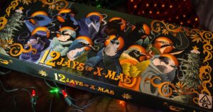 Festive dark green box titled '12 Jays of X-Mas,' adorned with colorful jays smoking cannabis joints, golden scrollwork, and cannabis leaves, set against a blurred background of Christmas lights, celebrating the holiday spirit with a cannabis twist