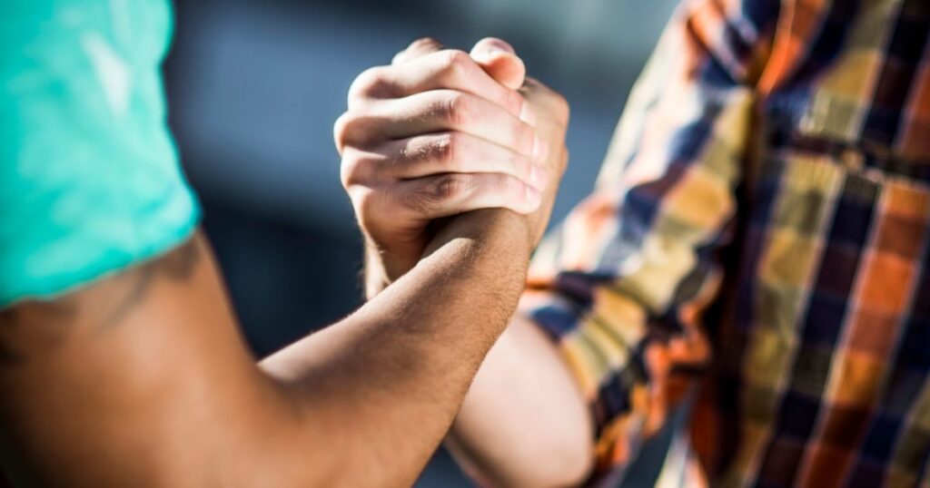 Close-up of two clasped hands in a firm grip, symbolizing solidarity and support, with blurred arms and casual clothing in the background, highlighting the theme of connection for International Men's Day.