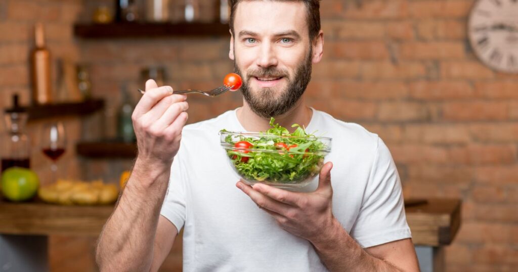 Bearded man in a white t-shirt holding a bowl of fresh salad and a fork with a cherry tomato, smiling warmly in a rustic kitchen setting, symbolizing health and wellness for International Men's Day