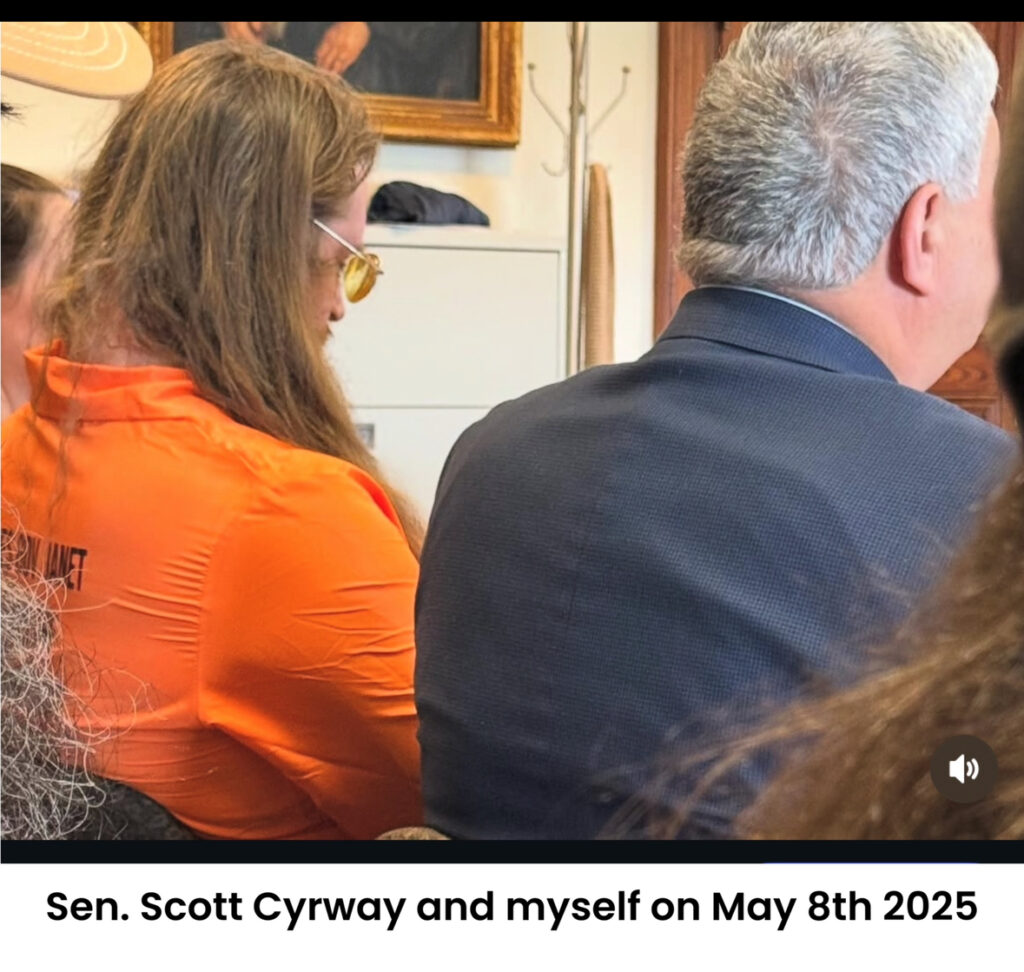 Senator Scott Cyrway in a dark blue suit and Derek Shirley in an orange shirt with 'SEN JANET' partially visible, captured from behind in an office setting with a filing cabinet, framed picture, and coat rack in the background, dated May 8th, 2025