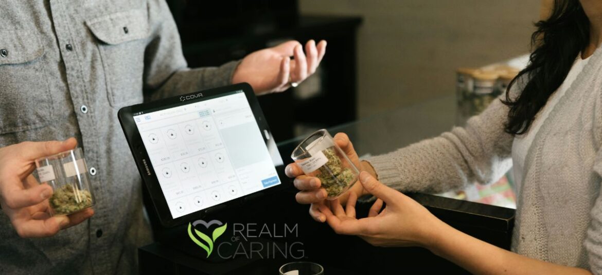 Retail transaction at a cannabis dispensary featuring a COVA point-of-sale system, glass containers with cannabis buds, and two individuals exchanging products, symbolizing Realm of Caring research advancements in cannabis science