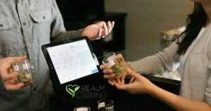 Retail transaction at a cannabis dispensary featuring a COVA point-of-sale system, glass containers with cannabis buds, and two individuals exchanging products, symbolizing Realm of Caring research advancements in cannabis science