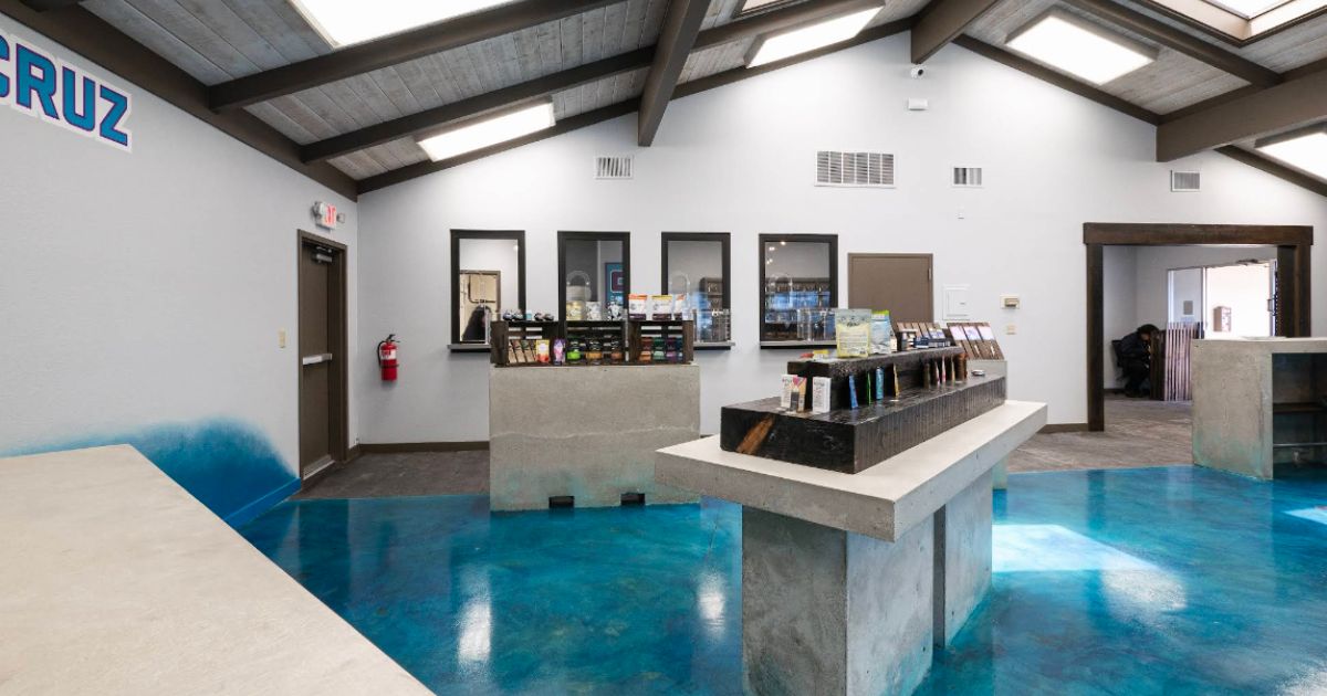 Interior of the CannaCruz dispensary in Salinas, featuring a vaulted wooden ceiling with skylights, vibrant blue reflective flooring, modern concrete display units, and neatly arranged cannabis products in a clean, industrial-chic retail space