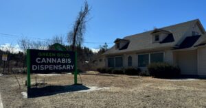 Exterior of Green Gold Cannabis Dispensary in Palmer, MA, featuring a prominent green and black sign, a light-colored building, and a clear blue sky, highlighting a potential location to find RSO near me