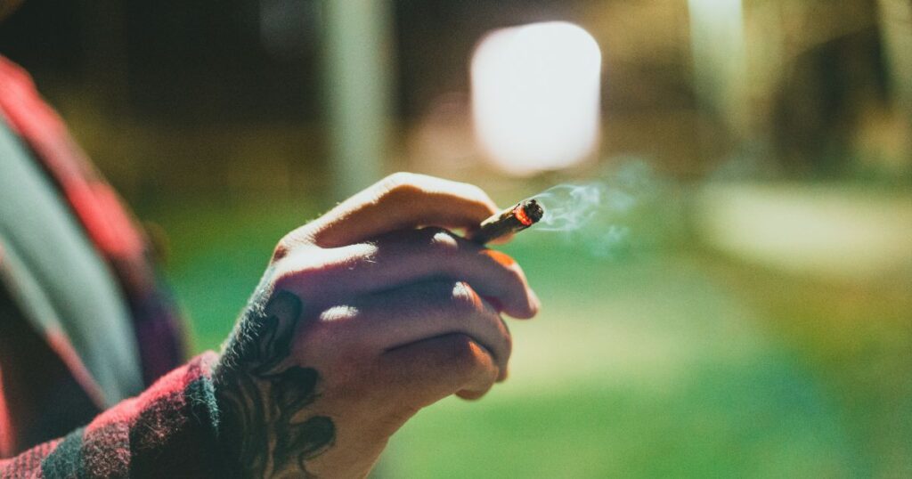 Close-up of a tattooed hand holding a lit smoking item with smoke curling upwards, set against a blurred outdoor background, symbolizing the 'California sober' lifestyle of substituting cannabis for alcohol