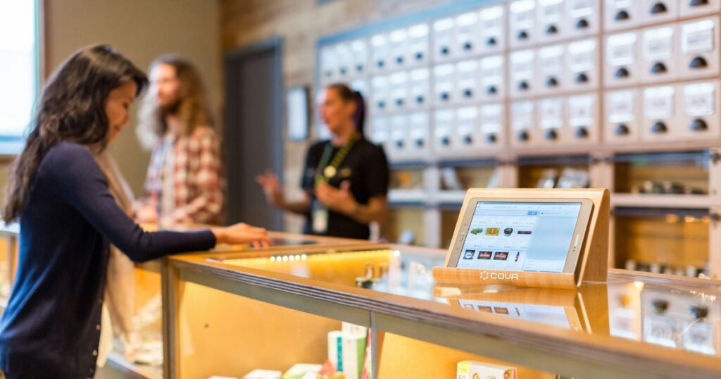 Customer interacting with a cannabis consultant at a modern dispensary, featuring a tablet displaying product options, glass display counters, and organized wooden product shelves, highlighting cannabis consultant best practices