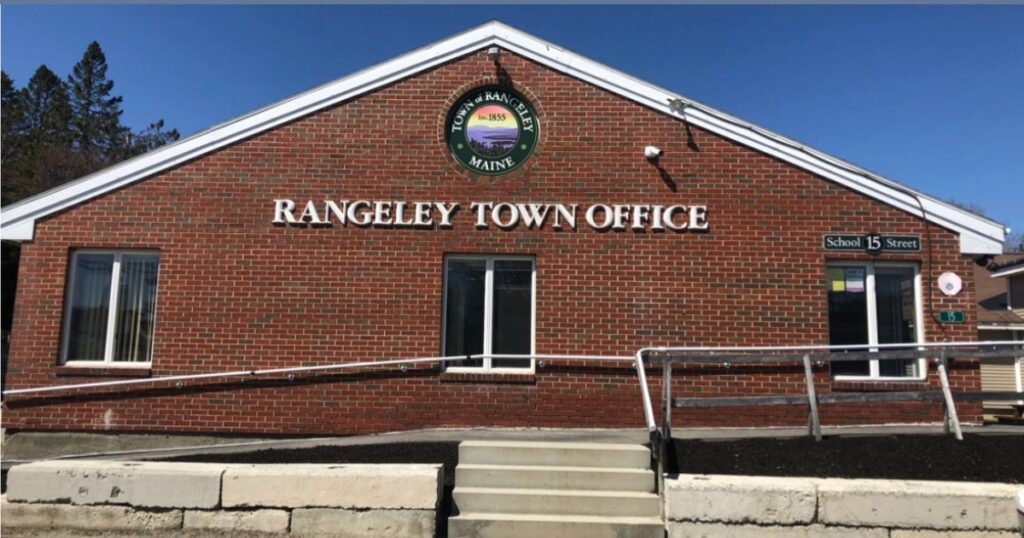 Front view of the Rangeley Town Office in Maine, a red brick building with white signage and a scenic town seal, set against a clear blue sky, symbolizing the backdrop of the Luke Sirois scandal