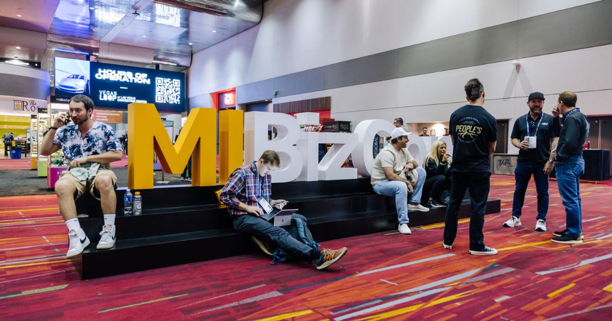 Indoor scene at MJBizCon featuring attendees networking, working on laptops, and making phone calls near large 'MiBizCo' branding letters, with a digital screen displaying event details, symbolizing the dynamic atmosphere of Media Day