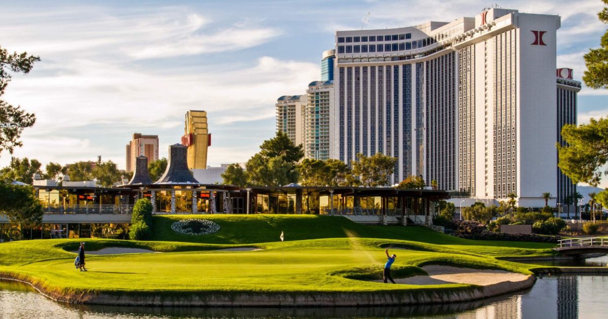 Scenic view of a golf course with a golfer mid-swing, a caddy walking nearby, and a clubhouse in the midground, set against tall hotel buildings under a bright sky, representing James Keyes’ Charity Driven Golf Classic.