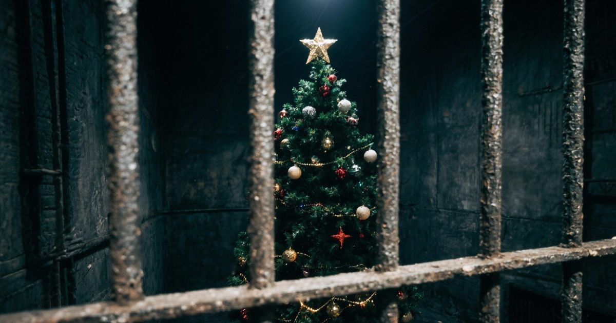 View of a decorated Christmas tree with a golden star topper, red and gold ornaments, and glowing lights, seen through rusty metal bars in a dimly lit room, symbolizing Edwin Rubis's experience of Christmas in prison