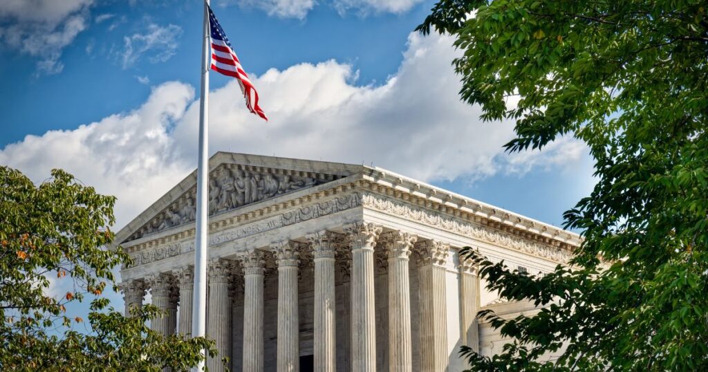 Photograph of the U.S. Supreme Court building (SCOTUS) with its neoclassical architecture, featuring tall Corinthian columns, a sculpted pediment, and the inscription 'JUSTICE UNDER LAW.' An American flag waves in the foreground under a bright blue sky with scattered clouds, framed by green foliage. The image symbolizes the solemnity and authority of the court, relevant to the ongoing marijuana case deliberations