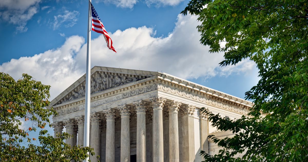 Photograph of the U.S. Supreme Court building (SCOTUS) with its neoclassical architecture, featuring tall Corinthian columns, a sculpted pediment, and the inscription 'JUSTICE UNDER LAW.' An American flag waves in the foreground under a bright blue sky with scattered clouds, framed by green foliage. The image symbolizes the solemnity and authority of the court, relevant to the ongoing marijuana case deliberations