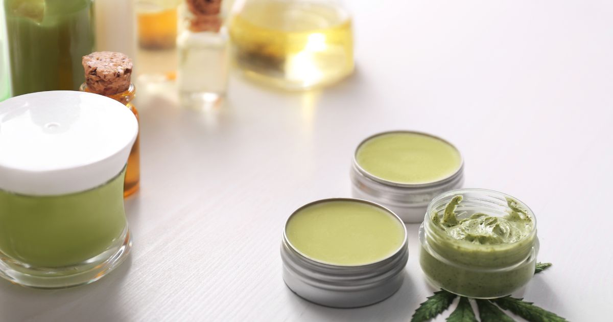 An arrangement of green cannabis-infused balms, creams, and oils in jars and tins, accompanied by natural green leaves, set against a clean white background, symbolizing cannabis as a potential relief for AIMSS symptoms