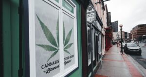 Street view of a storefront featuring a cannabis leaf graphic and text reading 'CANNABIS Licensed Retailer SOLD HERE,' set against a light green facade. The scene includes an urban street in Virginia with parked cars, streetlights, and other commercial buildings under an overcast sky, highlighting the presence of legal cannabis retail in the area.