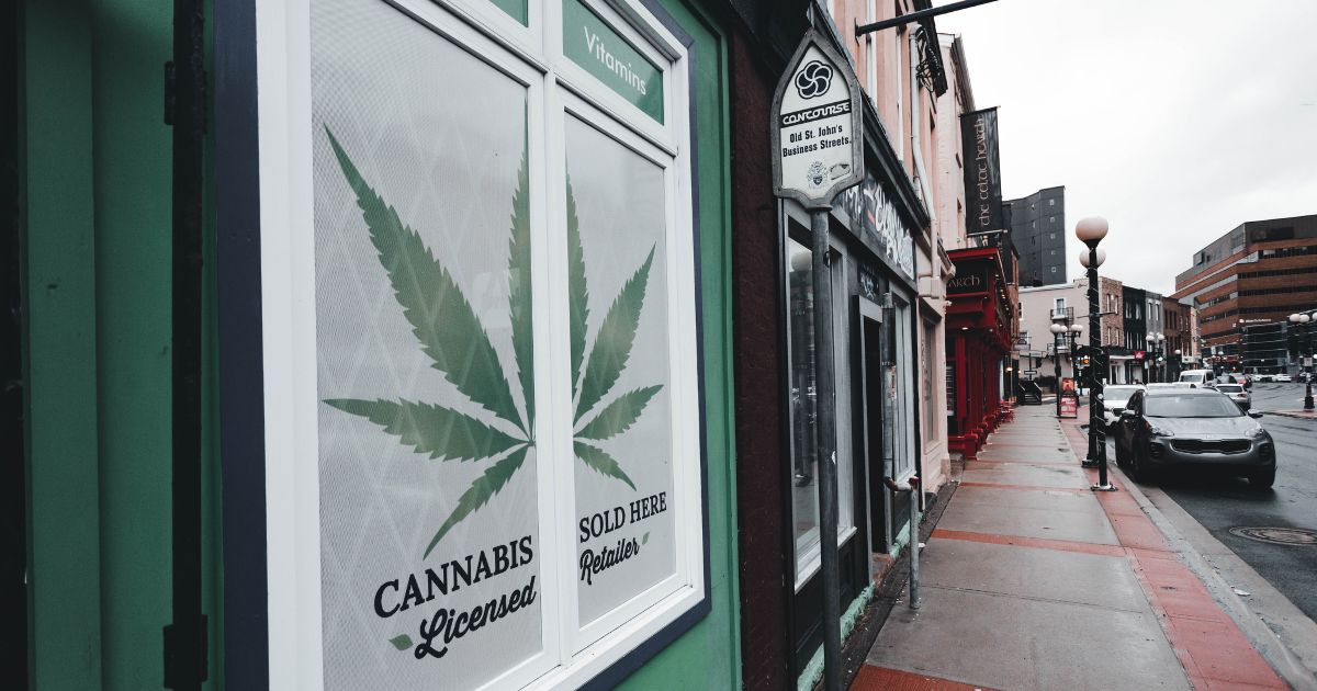Street view of a storefront featuring a cannabis leaf graphic and text reading 'CANNABIS Licensed Retailer SOLD HERE,' set against a light green facade. The scene includes an urban street in Virginia with parked cars, streetlights, and other commercial buildings under an overcast sky, highlighting the presence of legal cannabis retail in the area.