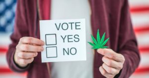 Close-up of hands holding a 'VOTE YES/NO' ballot and a green cannabis leaf, set against a blurred American flag background, symbolizing the Massachusetts adult-use cannabis repeal decision