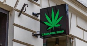 Close-up of a cannabis dispensary exterior featuring a black sign with a green cannabis leaf and 'CANNABIS SHOP' text, mounted on a light-colored building facade with decorative molding.