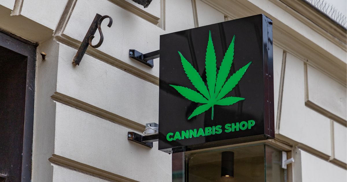 Close-up of a cannabis dispensary exterior featuring a black sign with a green cannabis leaf and 'CANNABIS SHOP' text, mounted on a light-colored building facade with decorative molding.