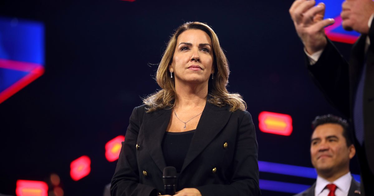 A woman, identified as Sara Carter, holding a microphone and gazing upward on a dimly lit stage with abstract red and blue lights in the background, symbolizing her role as the new Drug Czar