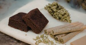 Still life featuring cannabis brownies, dried buds, pre-rolled joints, and loose ground cannabis on a textured fabric, symbolizing different consumption methods and their varying cannabis effects