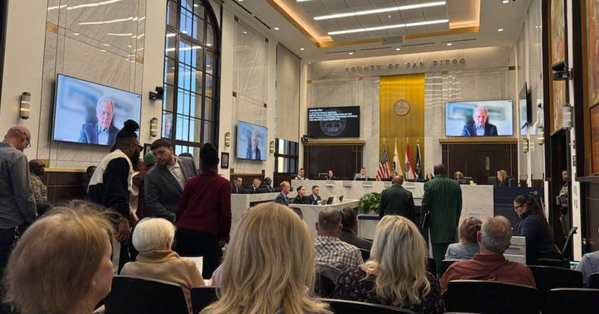 Formal government meeting in San Diego County with officials seated at a dais, audience members in attendance, and a remote participant displayed on large screens, symbolizing discussions on the social equitable cannabis program.