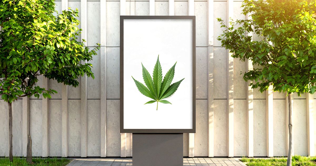 Outdoor scene featuring a vertical billboard with a vibrant green cannabis leaf on a white background, flanked by two green trees against a modern concrete wall, symbolizing innovative cannabis marketing strategies.