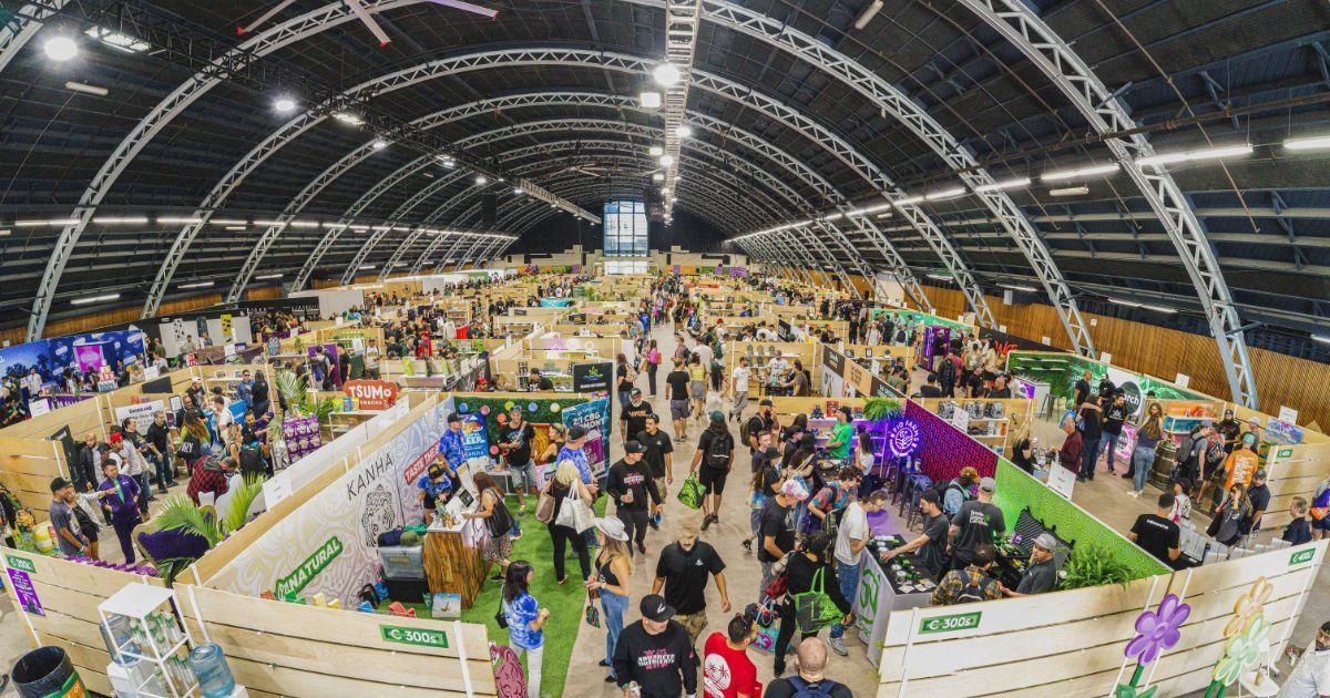 Wide-angle view of a bustling indoor cannabis event with rows of wooden booths showcasing brands like 'TSUMO SNACKS' and 'KANHA.' Attendees explore displays featuring cannabis products, greenery, and vibrant branding under a large arched ceiling, highlighting the energy of cannabis trade shows.
