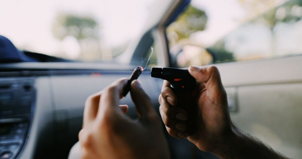 Close-up of hands inside a car, one holding a lit blunt with smoke rising and the other using a torch lighter, symbolizing the context of California's Supreme Court cannabis ruling on how police handle cannabis in vehicles