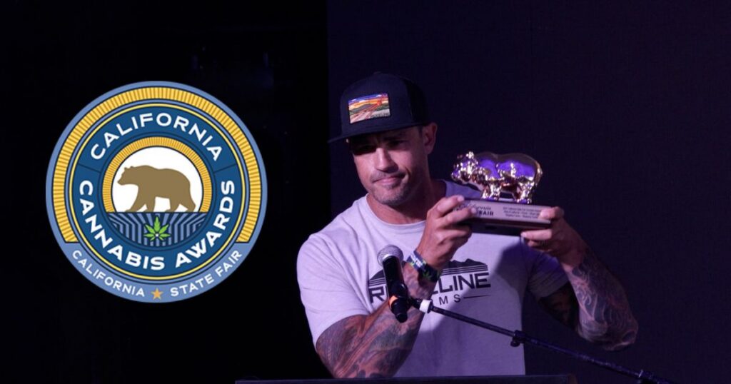 Image of a man holding a golden trophy featuring two bear figures at the California Cannabis Awards 2026. He wears a 'Ridgeline Farms' t-shirt and a black cap with a landscape patch, standing behind a microphone. A circular logo for the California Cannabis Awards and State Fair, with a bear and cannabis leaf, is prominently displayed on the left against a dark event backdrop