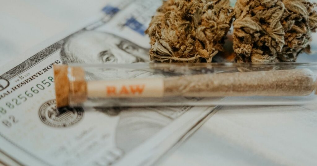 Close-up of US hundred-dollar bills serving as a backdrop for two dried cannabis buds and a pre-rolled joint in a clear tube labeled 'RAW.' The composition symbolizes the financial and transactional aspects of cannabis banking