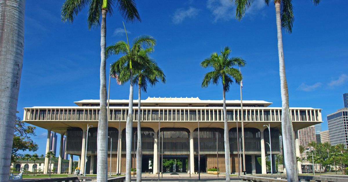 Wide shot of a modern government building in Hawaii, framed by tall palm trees under a vibrant blue sky. The building's unique architecture, with overhanging sections and vertical columns, symbolizes the legislative setting for Hawaii's cannabis bill discussions