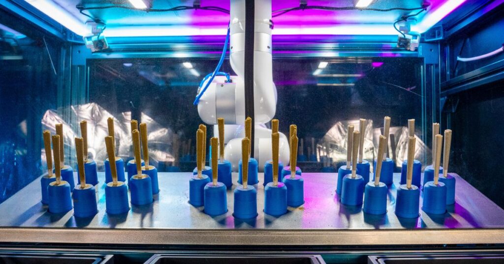 Close-up of an automated system featuring a white robotic arm positioned above rows of pre-rolled cannabis cones held upright in blue holders. The scene is illuminated by vibrant blue and purple lights, highlighting the advanced technology used in the cannabis industry for precision and efficiency