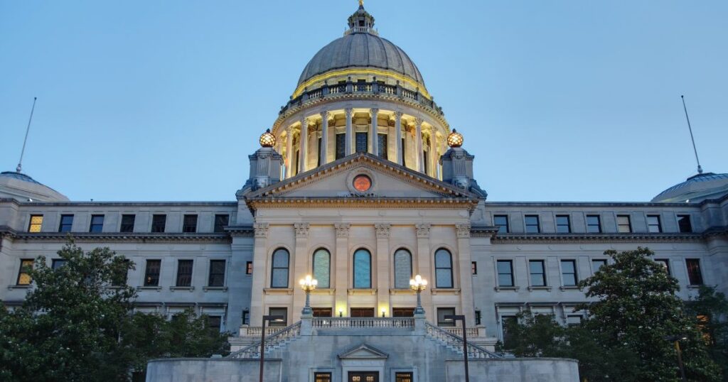 Illuminated neoclassical government building with a grand dome and symmetrical wings, set against a clear blue sky. The structure symbolizes legislative progress, referencing Mississippi's Ryan's Law for terminally ill patients.