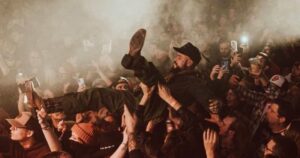 Dynamic concert scene with Edward Crowe being crowd-surfed above a dense, energetic audience. The smoky atmosphere, illuminated by smartphone lights, captures the vibrant energy of the event, symbolizing the intersection of music, community, and psychedelics