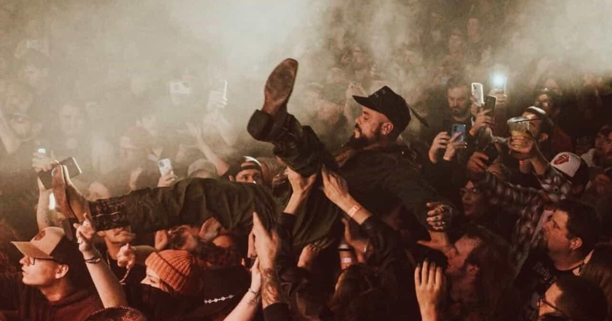 Dynamic concert scene with Edward Crowe being crowd-surfed above a dense, energetic audience. The smoky atmosphere, illuminated by smartphone lights, captures the vibrant energy of the event, symbolizing the intersection of music, community, and psychedelics