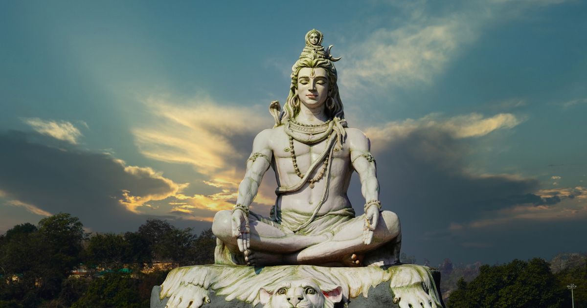 Majestic statue of Lord Shiva seated in a meditative pose, adorned with a crescent moon, a cobra on his shoulder, and a third eye on his forehead. The statue is set against a dramatic sky with hues of blue, gray, and orange, symbolizing the spiritual essence of Maha Shivaratri. Dark green trees frame the base, enhancing the serene and sacred atmosphere