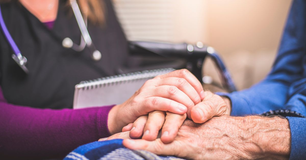 Close-up of a caregiver gently holding the hand of an elderly person, symbolizing compassion and support. A stethoscope and wheelchair are visible in the softly blurred background, representing a healthcare setting. The image relates to Ryan's Law in Oregon, focusing on medical cannabis for hospice patients