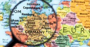 Close-up of a political map of Europe with a magnifying glass centered on Germany, highlighting cities like Berlin, Munich, and Hamburg. The image symbolizes the focus on Germany's cannabis industry and the Organigram-Sanity Group acquisition, emphasizing the region's growing significance in the cannabis market