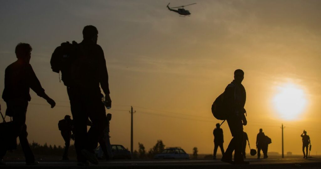 Silhouetted group of individuals, possibly veterans, walking along a road at sunrise or sunset, carrying backpacks and bags. A helicopter flies in the background against a warm, gradient sky. The scene symbolizes the journey toward equitable access to medical cannabis for veterans