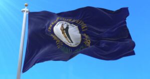 The Kentucky state flag waves against a clear blue sky, featuring a dark blue field with the state seal at its center. The seal shows two figures shaking hands, symbolizing unity, surrounded by the motto 'United We Stand, Divided We Fall,' representing Kentucky's connection to cannabis legislation efforts