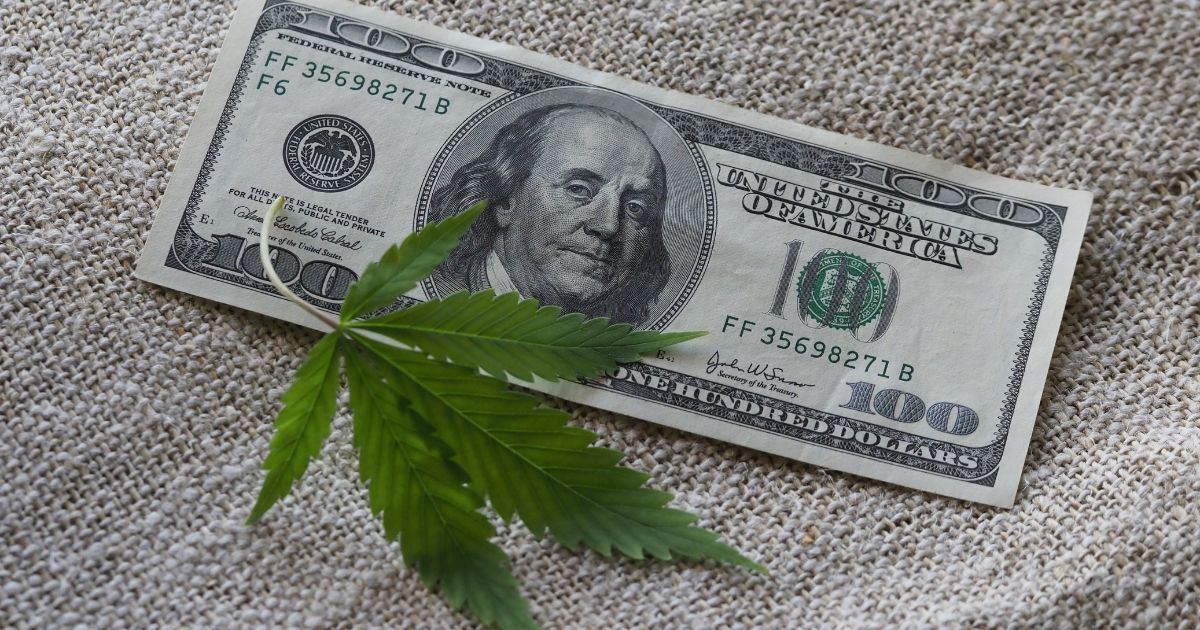 Close-up of a one-hundred dollar bill partially covered by a green cannabis leaf, placed on a textured beige fabric. The image symbolizes the financial impact of Michigan's cannabis tax