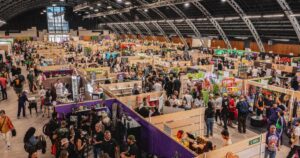 Wide-angle view of a bustling indoor trade show featuring vendor booths, branded banners, and a diverse crowd engaging with exhibits, highlighting the impact of cannabis event sponsorship strategies in a vibrant industry setting.