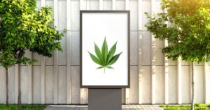 Outdoor billboard featuring a vibrant green cannabis leaf on a white background, flanked by small trees and set against a modern building facade. The clean, minimalist design highlights a cannabis marketing strategy focused on simplicity and visibility