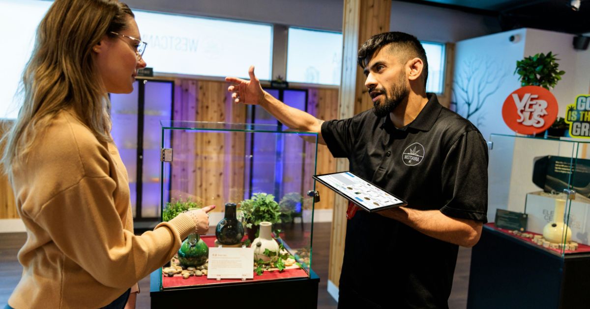 Modern cannabis dispensary scene featuring a staff member in a branded polo shirt using a tablet to assist a customer pointing at a glass display case with curated products, symbolizing engagement and ownership in retail operations.