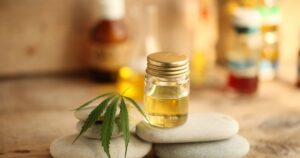 Close-up of a small clear glass bottle filled with golden-yellow liquid, likely CBD oil, with a golden screw-top lid. The bottle rests on a stack of smooth, light-colored stones, accompanied by a vibrant green cannabis leaf. The blurred background features additional bottles and a wooden surface, evoking a natural or wellness-focused setting. Keywords: CBD study.