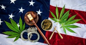 Flat lay composition featuring an American flag as the background, with a wooden gavel, silver handcuffs, green cannabis leaves, a bowl of dried cannabis flower, and two rolled joints arranged on top. The image symbolizes the intersection of cannabis and legal or governmental policies. Keywords: Army eases cannabis rules