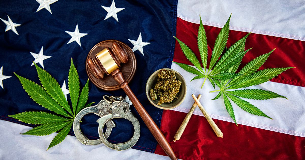 Flat lay composition featuring an American flag as the background, with a wooden gavel, silver handcuffs, green cannabis leaves, a bowl of dried cannabis flower, and two rolled joints arranged on top. The image symbolizes the intersection of cannabis and legal or governmental policies. Keywords: Army eases cannabis rules